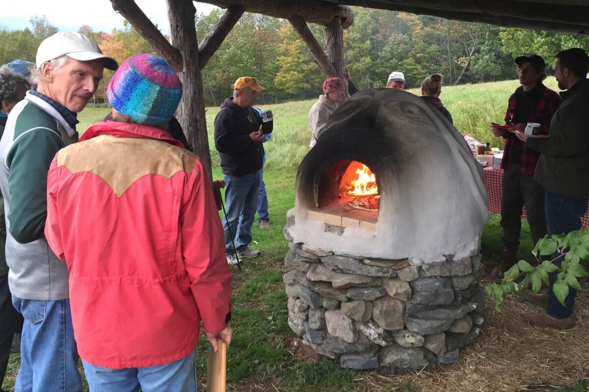 People gathered around wood-fired earth oven, talking.