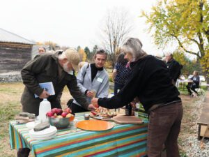 Three people stand around a table of food outdoors, with several other people milling the background.