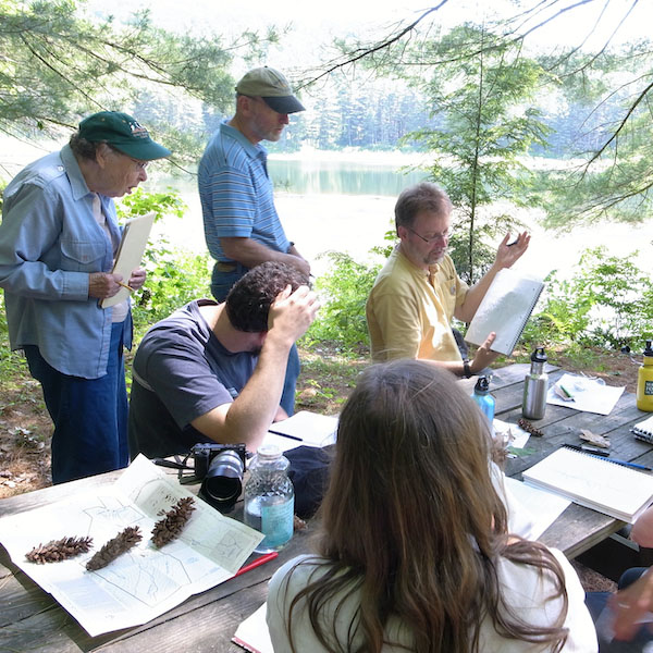 Students listen to their instructor at a picnic table on the bank of a pond